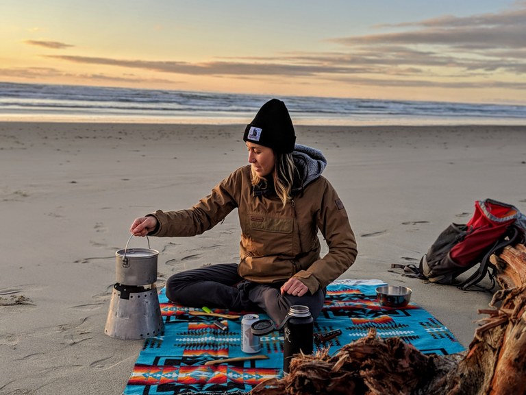 A person sitting on a blanket on the beach, heating up water in a camp stove. 
