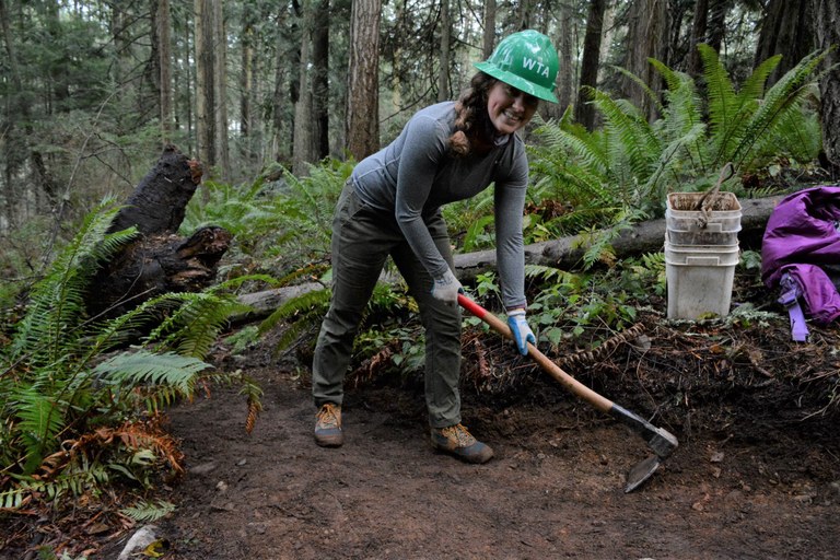 WTA staffer Rebecca stands on trail and smiles at the camera while using a grub how to flatten out the trail tread.