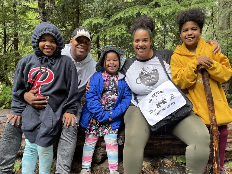 Two parents and two kids sit on a log in the forest wearing campfire coffee cloting.. 
