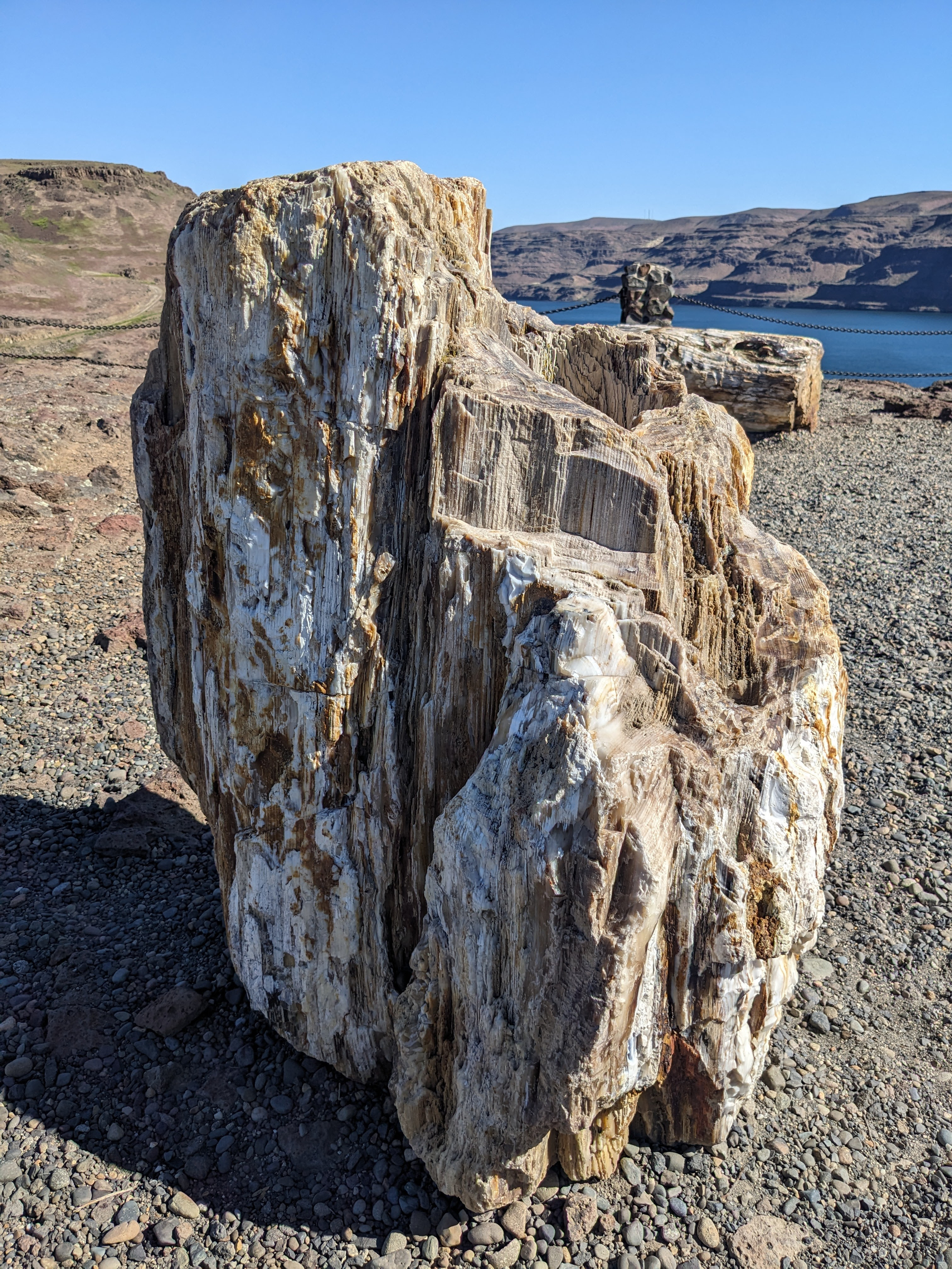 Large rock stands out amongst smaller pebbles that make up the ground. Blue skies, rolling hills and rivers seen in the background.