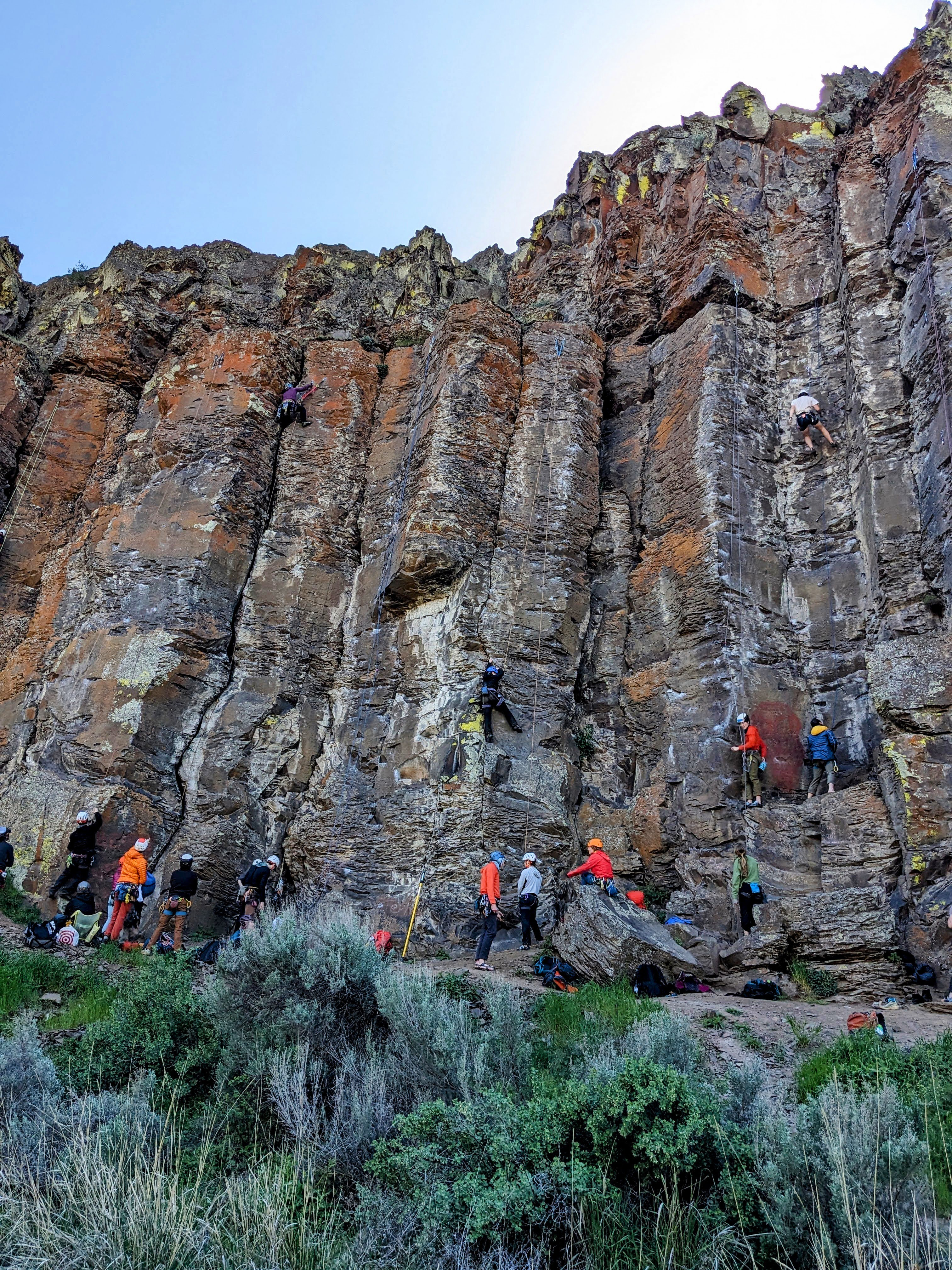 10 climbers at the bottom of rock face amoungst shrubs and boulders. Three climbers are on various spots on the wall. 