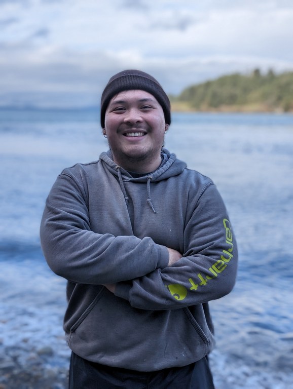 Riley by the sea at an Emerging Leaders Program work party. Photo by Tiffany Chou Riley poses with their arms crossed in front of the Salish Sea.