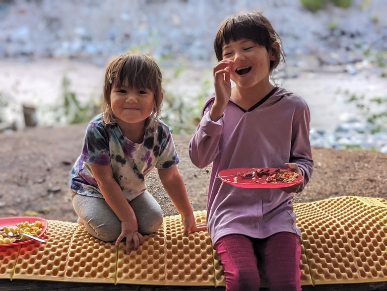 Two young sisters sit together near a river, enjoying a picnic. 
