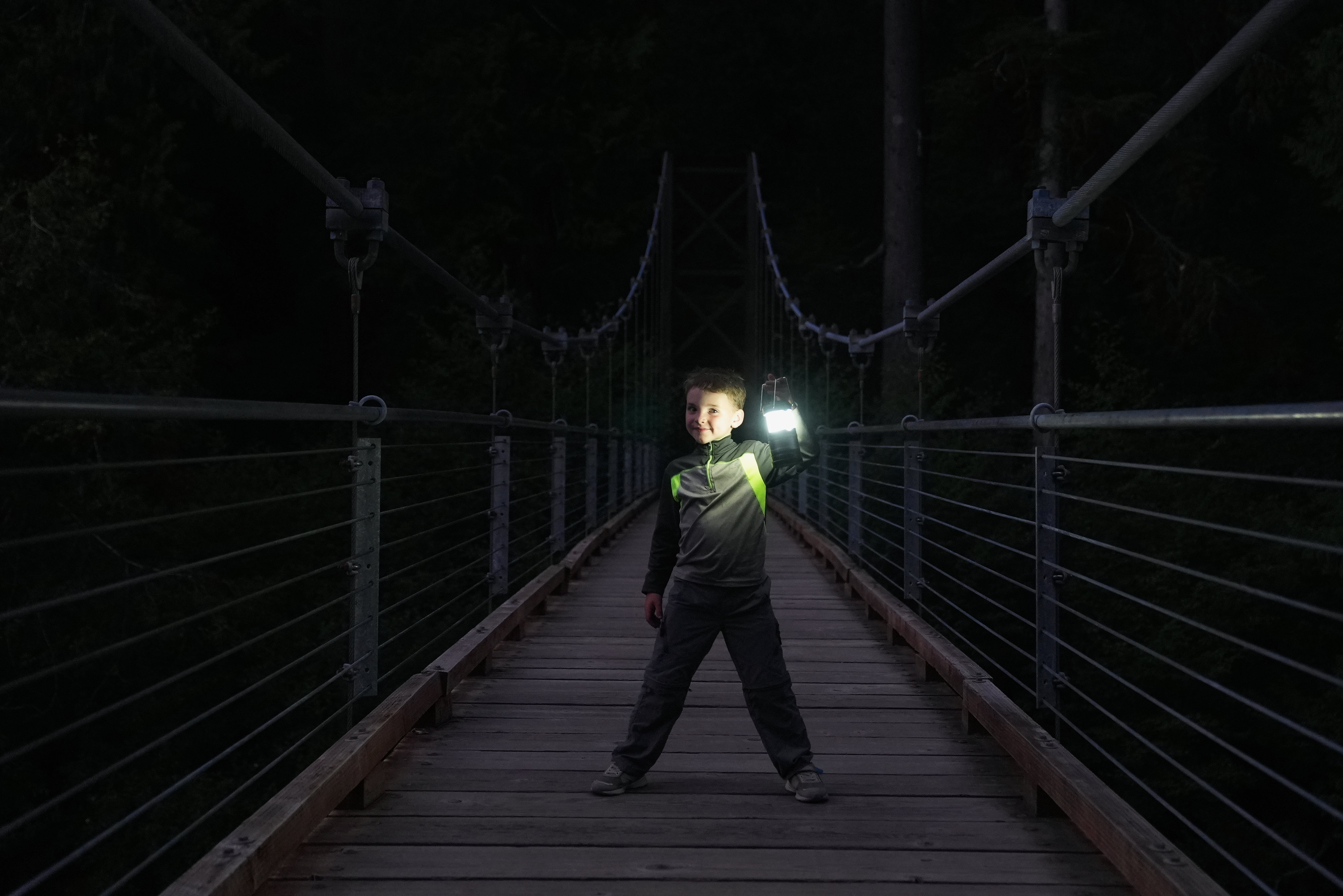 Child holds glowing lantern while standing on a bridge at dusk.