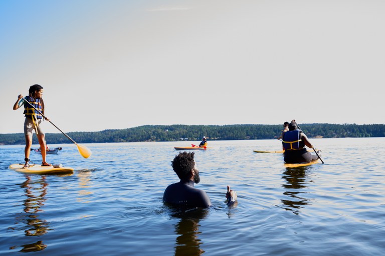 Five kayakers and stand up paddle-boarders paddle on a lake. One person stands in water watching.
