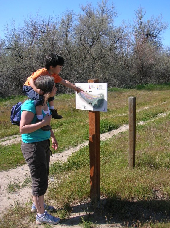 A child sits on a hikers shoulders and points at a trail sign.