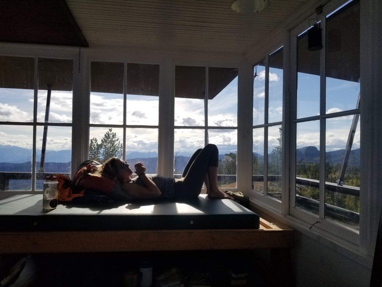 A hiker rests on a bed in a fire lookout.