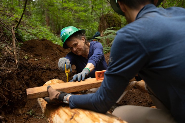 Volunteers work on installing deck boards on a brand new bridge at Wallace Falls State Park. Photo by Kesia “Lee” Lee. Two people work together on trail to measure wood wearing green hats.