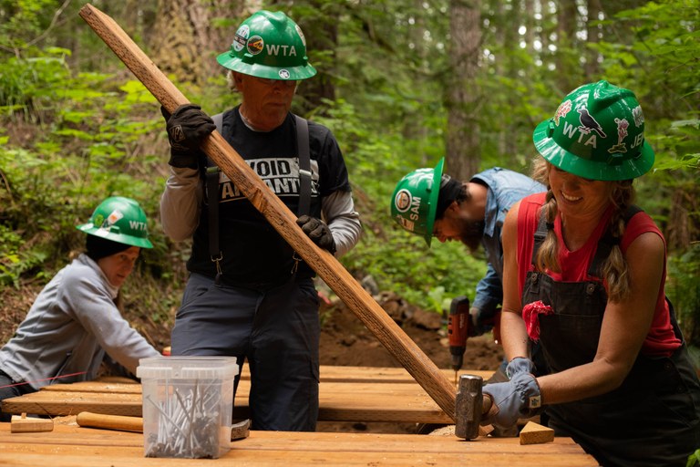 Volunteers work together to attach the last few deck boards to complete the bridge. Photo by Kesia “Lee” Lee. Four volunteers wearing green hats align pieces of wood while on trail.