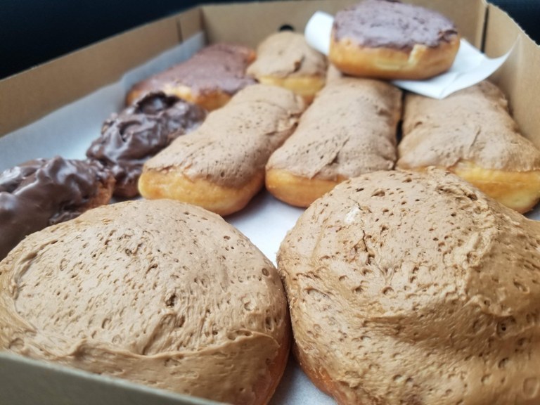 A dozen donuts sit in a box. The frosting on the maple bars seems particularly appealing.
