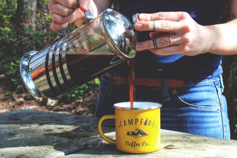A person pours coffee from a French press into a Campfire Coffee Co. mug