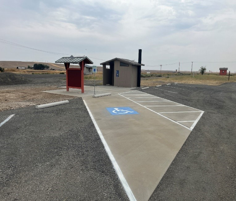 Parking lot with pit toilet and trailhead sign. Facing handicap parking space. rolling grass hill and cloudy skies in background.