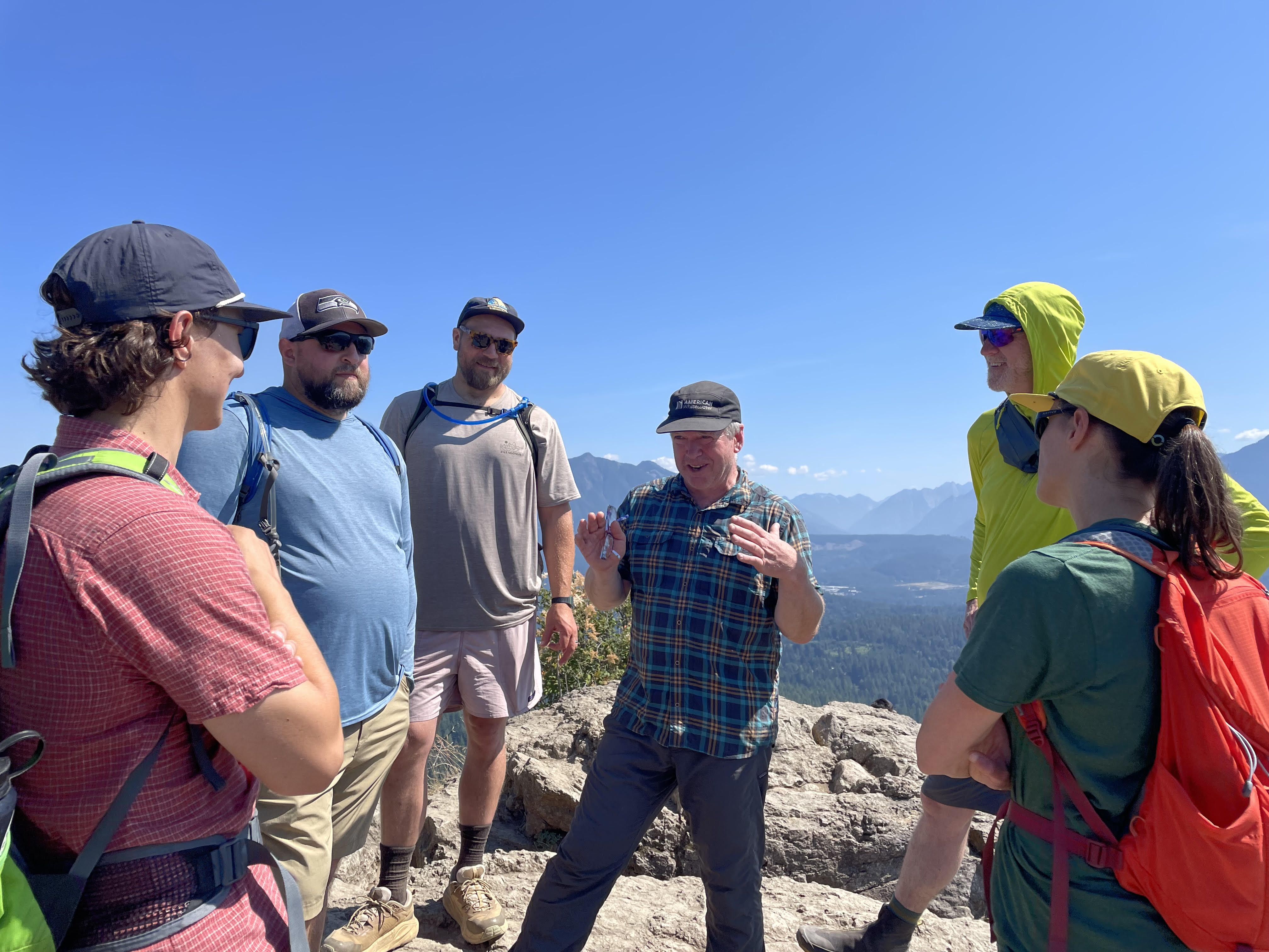 A group stands talking with mountain views behind them on bright, sunny day