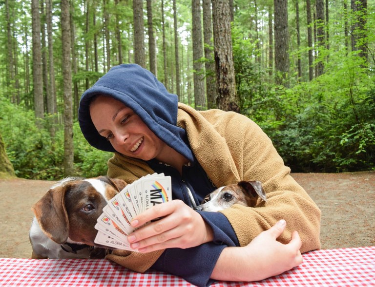A camper plays cards on a picnic table. They show their hand to their dog, who looks approving.