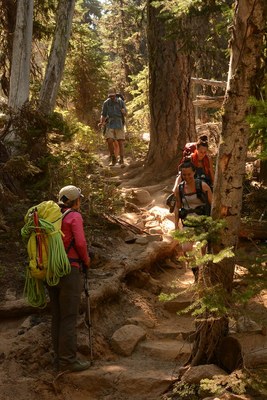 A hiker with a climbing rope stands to the side as 4 other hikers come downhill to pass. 
