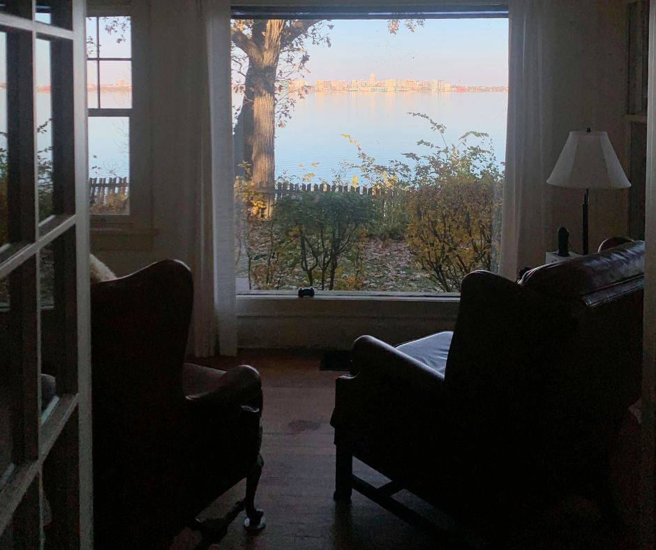 Lake House View by Victoria Obermeyer View of lake out of window on sunny day. City in background. Leather chairs and wood floor look out window over the water.