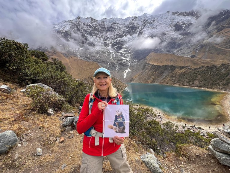 Woman in red jacket holding 8x11 print out of Miles the Marmot in front of blue, cloudy alpine lake.
