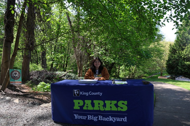 Person smiles at table in front of trail entry on sunny, green day. 