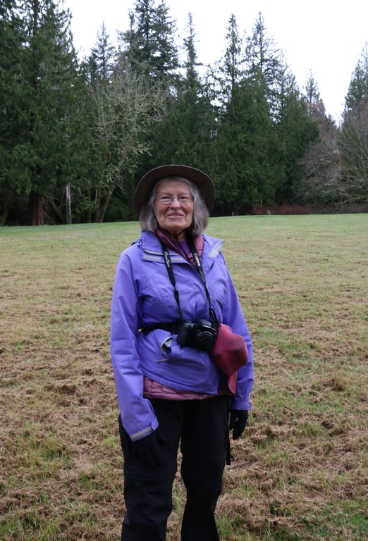 Women in purple coat stands in field with evergreens in background.