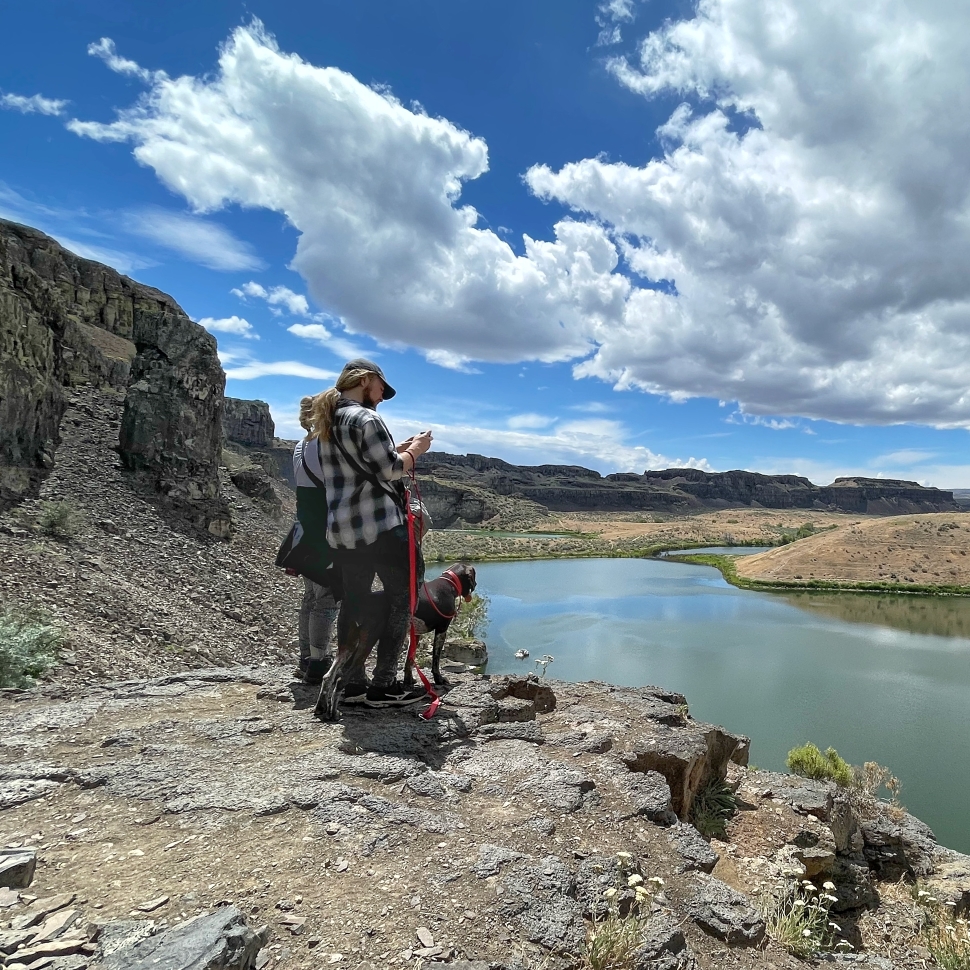 Ancient Lakes. Photo by Leap. Two adults and a leashed dog standing at the edge of the rocks taking a photo of the water at Ancient Lakes. Photo by Leap.