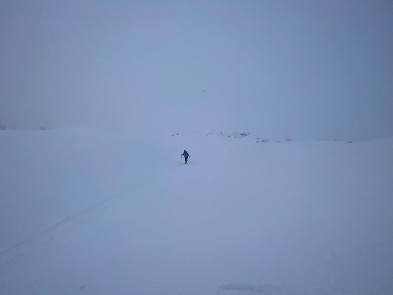A blue-jacketed hiker walks across a snowfield on a cloudy day 