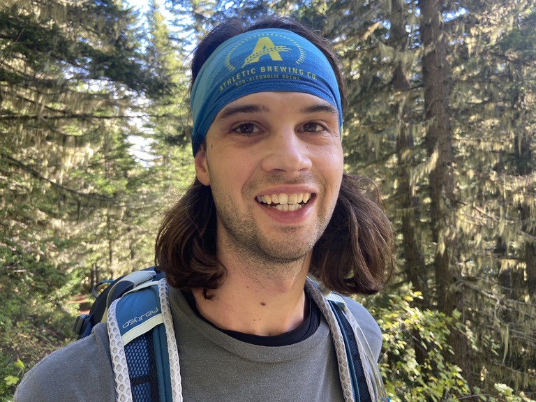 A hiker wearing a backpack and blue headband saying "Athletic Brewing" smiles at the camera, with evergreen trees in the background.