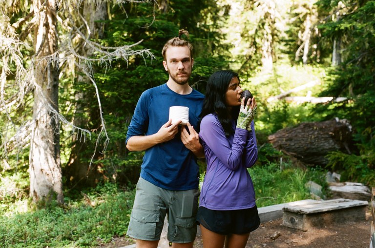 Two backpackers stand together. One is seriously holding a role of toilet paper. Another is kissing an avocado. 