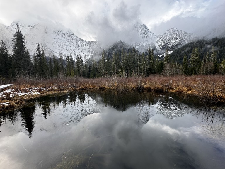 A reflective lake on the Big Four Ice Caves trail with snow-capped mountains in the background on a foggy day. Photo by trip reporter Shellyf.