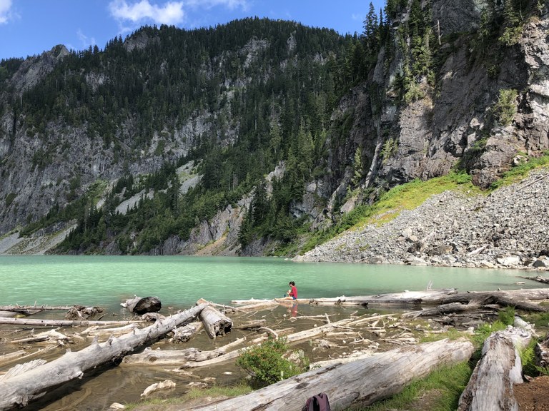 Blanca Lake A hiker sits on a log floating at the edge of a bright turquoise mountain lake.