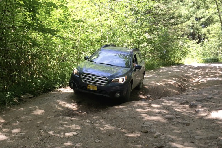 A car navigates a badly rutted road in green forest. The car is at an angle, and likely scraping its bottom on the ground. 