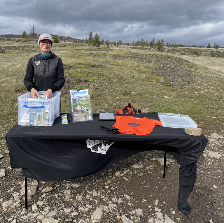 Ambassador poses behind an outreach table at the Catherine Creek Trailhead. The tablecloth is blowing in the wind.