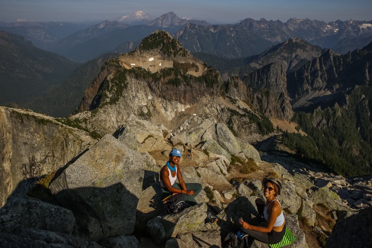 Two women of color sit on rocks, smiling up at the camera, with peaks reaching far into the distance. 