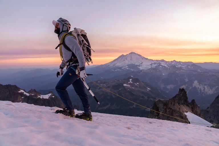  Outdoor Research ambassador Max Djenohan climbs a snow-covered peak as the sun sets over the mountains in the background.