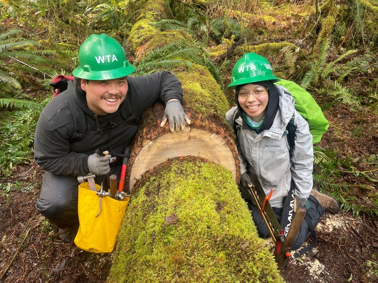 Riley and Valeria smiling at the camera next to a large cut log. Photo by Michelle Mouw.