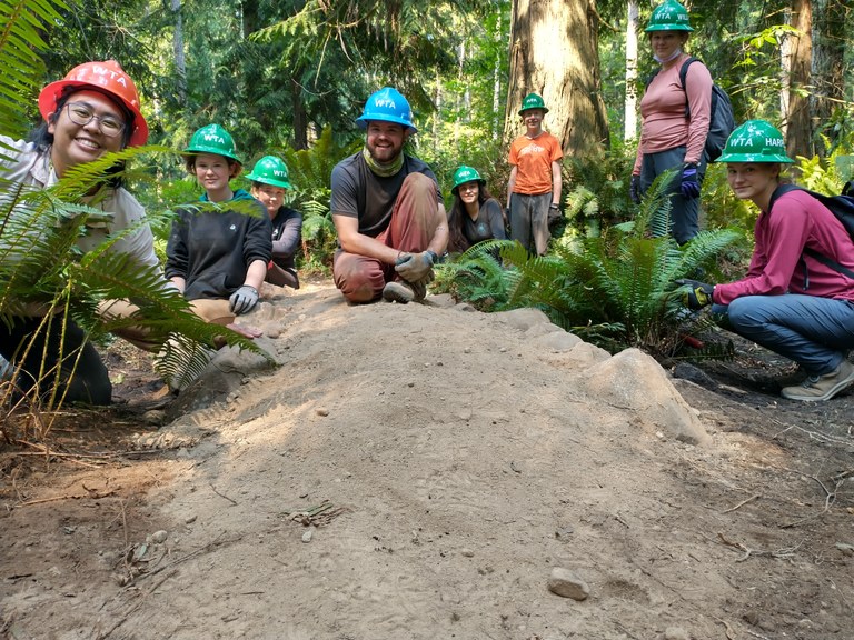 A groups of youth volunteers and leaders, wearing hard hats, smiles at the camera from behind stretch of fresh dirt surrounded by ferns.