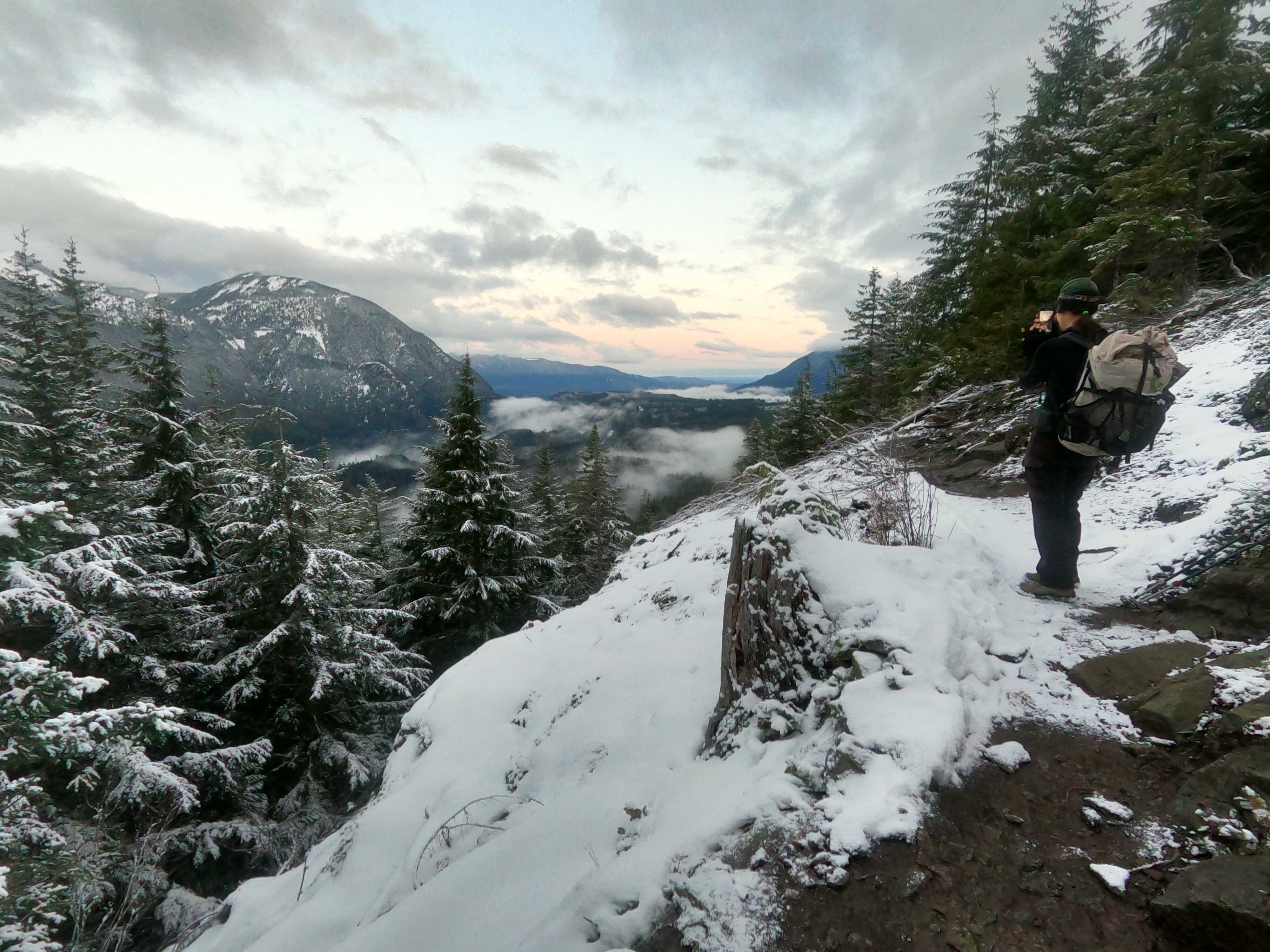 Dirty Harry's Balcony by Alex Kalaw Hiker stands on a snowy trail and takes a photo of the surrounding trees, rolling mountains, pastel sky