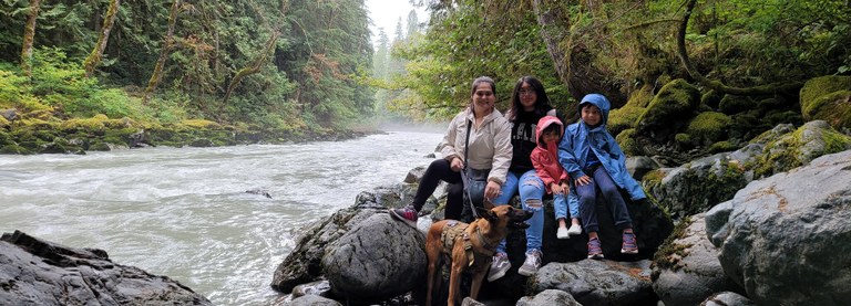 An adult hiker and three children sit on a boulder next to a flowing river, with lots of green evergreens in the background. The adult is holding a dog on a leash. Everyone is smiling, even the dog.