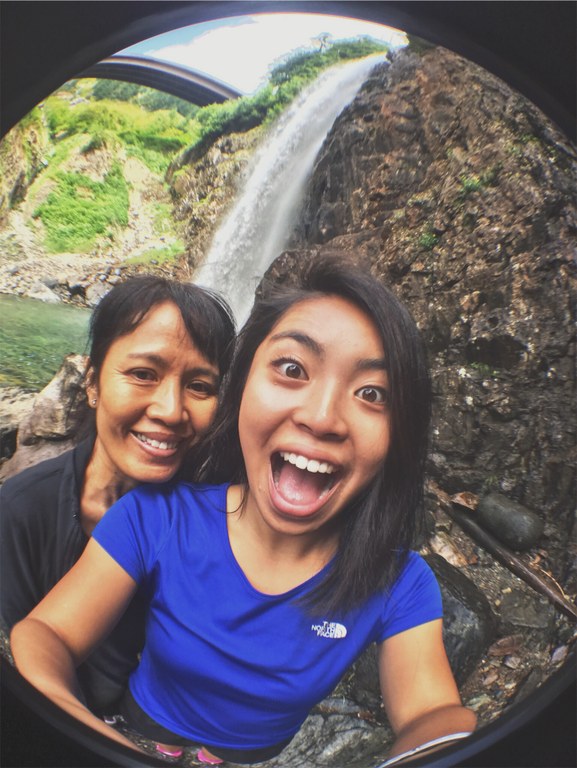 Two people with bright smiles pose for a selfie with a waterfall behind them.