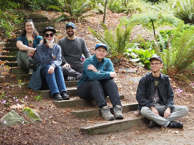Five people sit on an outdoor staircase in a garden, facing the camera and smiling. 