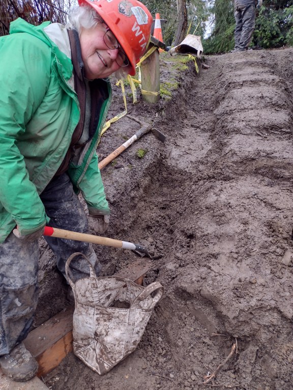 A volunteer wearing an orange hard hat and green jacket, works in the mud to help build new stairs at a park.