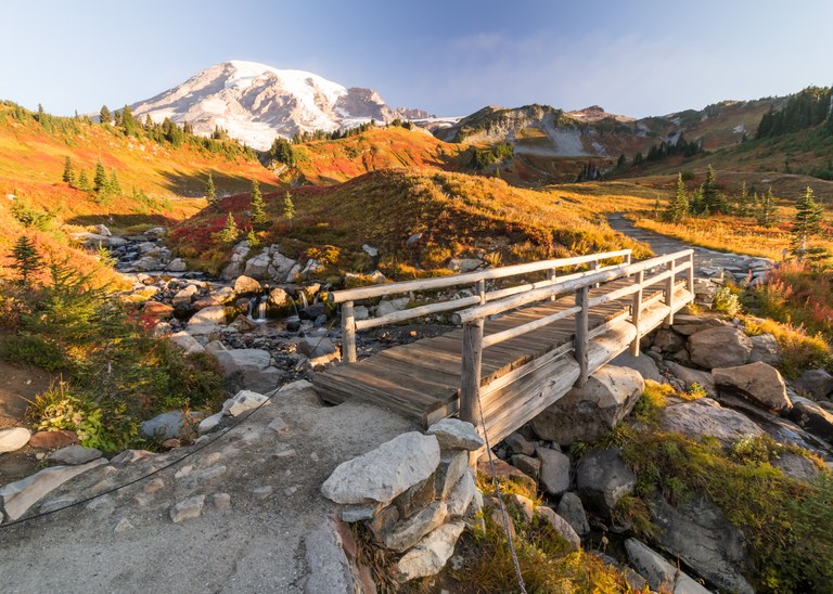 A wooden bridge spans a creek through a golden meadow with Mount Rainier in the distance. Photo by Muhamad Said. 