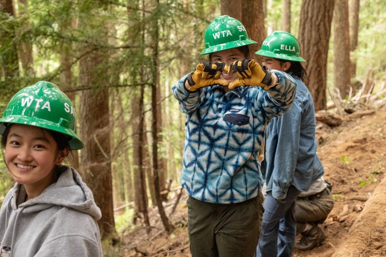 Three young female volunteers wearing hard hats and coats smile at the camera while one makes a heart with her fingers 