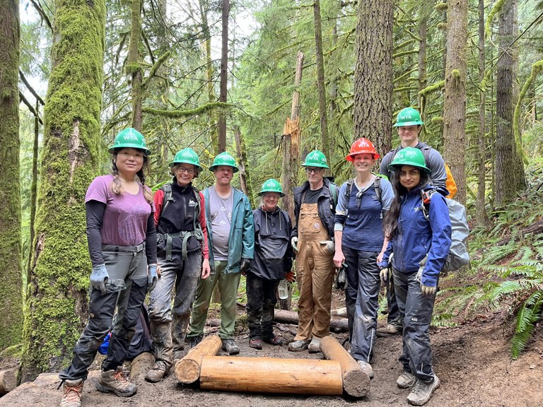 A group of volunteers wearing hard hats and gloves stand near a newly-built structure on trail. 