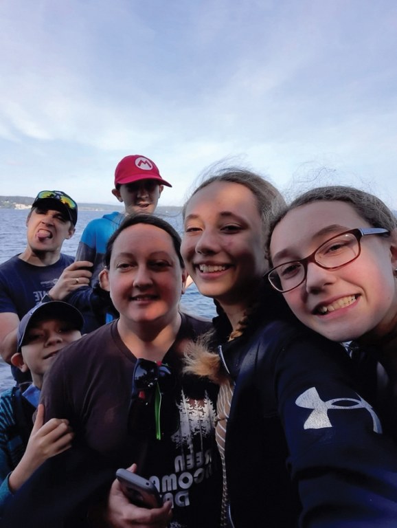 A family of six smiles for a selfie with blue water and blue sky in the background. 