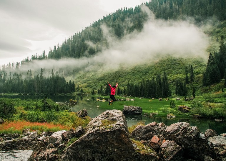 A hiker jumping in joy on top of a large boulder near a mountain lake.