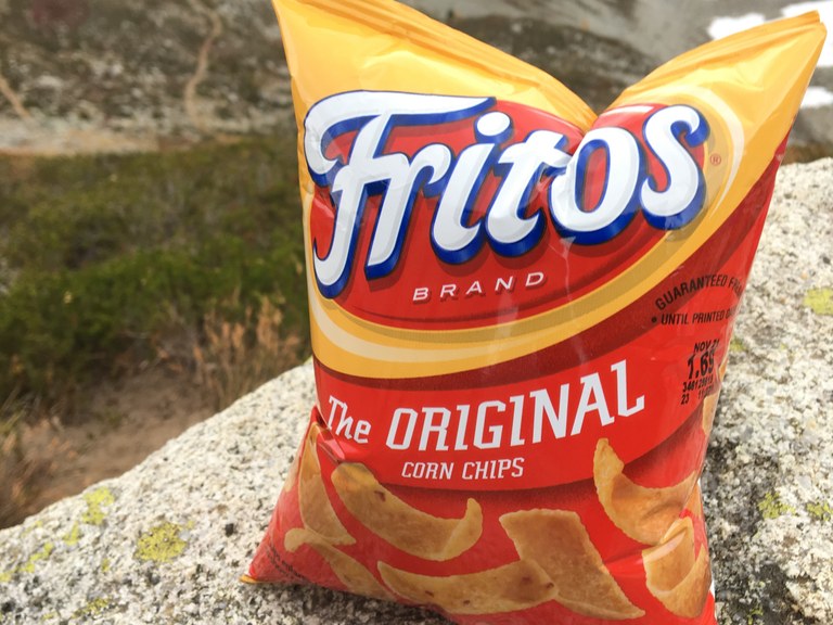 A large bag of Fritos rests on a rock above a green valley. 
