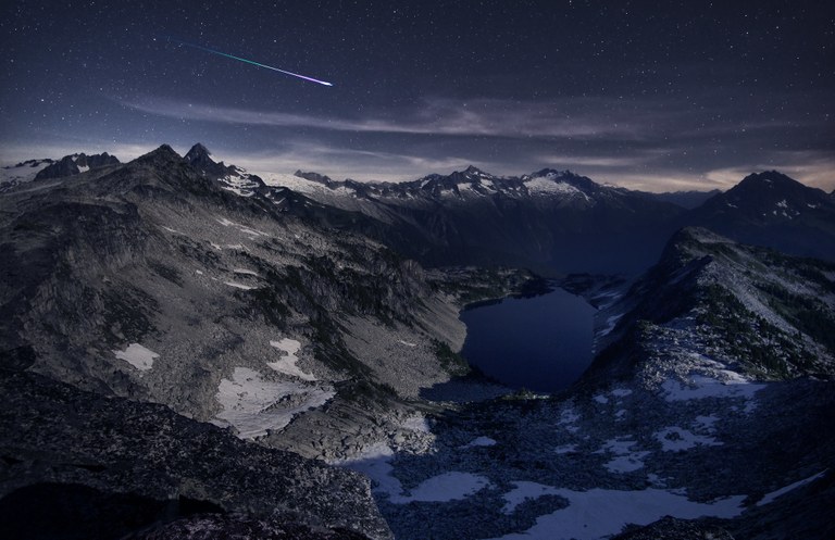 A meteor streaks across the sky over mountains and a lake.