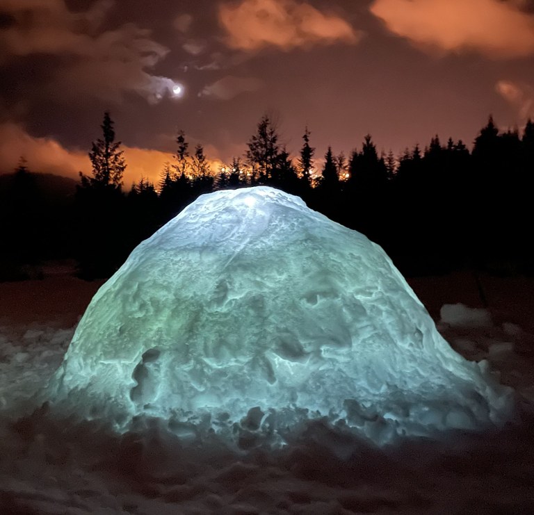 An igloo, lit from inside, glows green in front of a dark orange sunset. 
