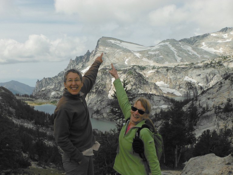 Enjoying backcountry views with WTA staff! Hikers stand in a mountain setting. They have their hands raised, pointing towards a peak in the background.
