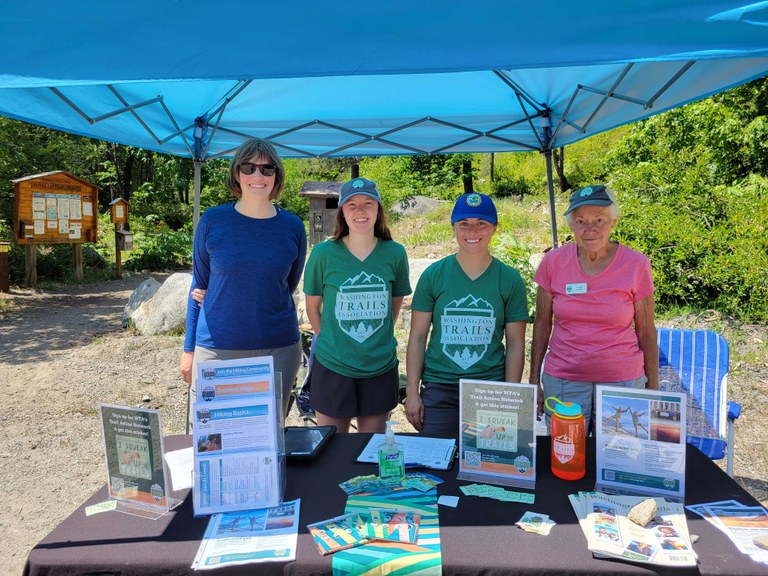 Four women, WTA staff and volunteers, pose behind a WTA outreach table at the Ingalls Creek Trailhead
